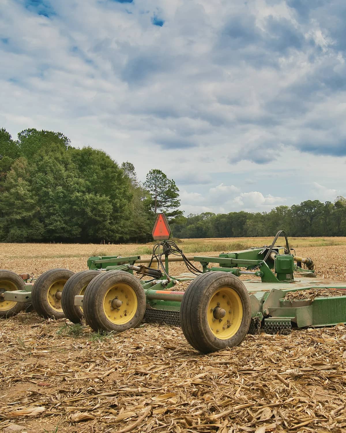Large batwing rotary cutter resting in a cut corn field, with a tree line behind.