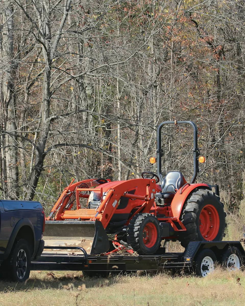 Pickup truck towing an orange compact tractor with a front loader on a flatbed trailer, beside a stand of bare autumn trees.