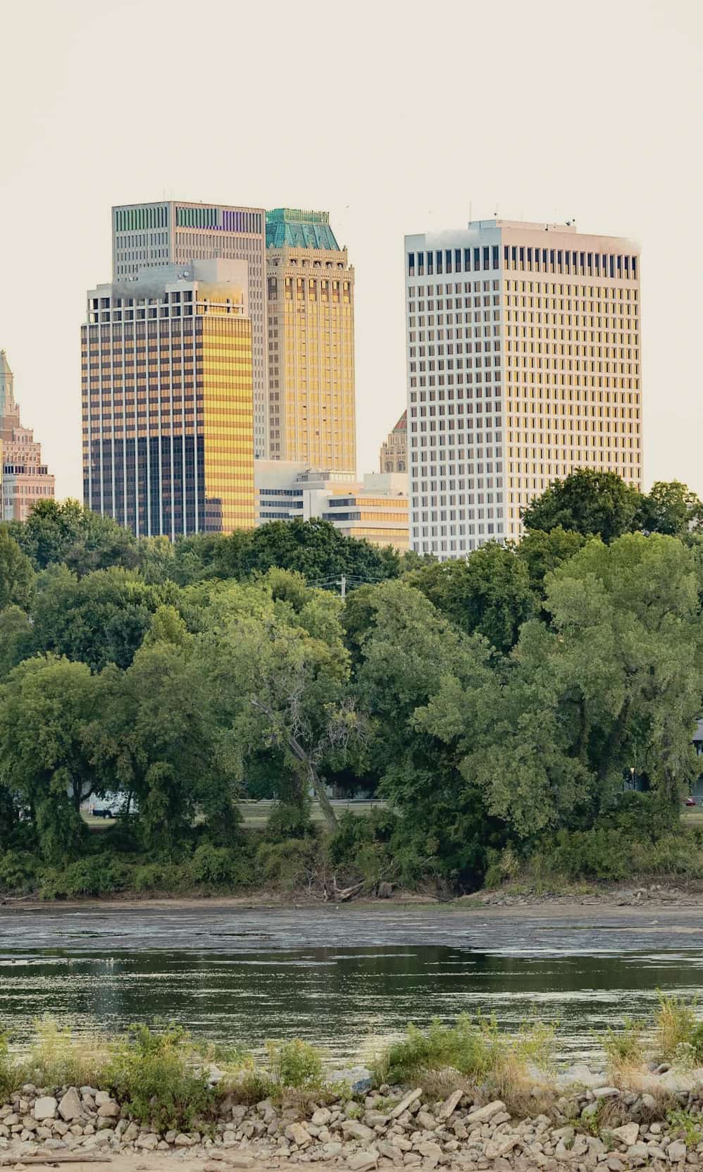 Downtown Tulsa towers in late-afternoon sun above a tree-lined bank of the Arkansas River.